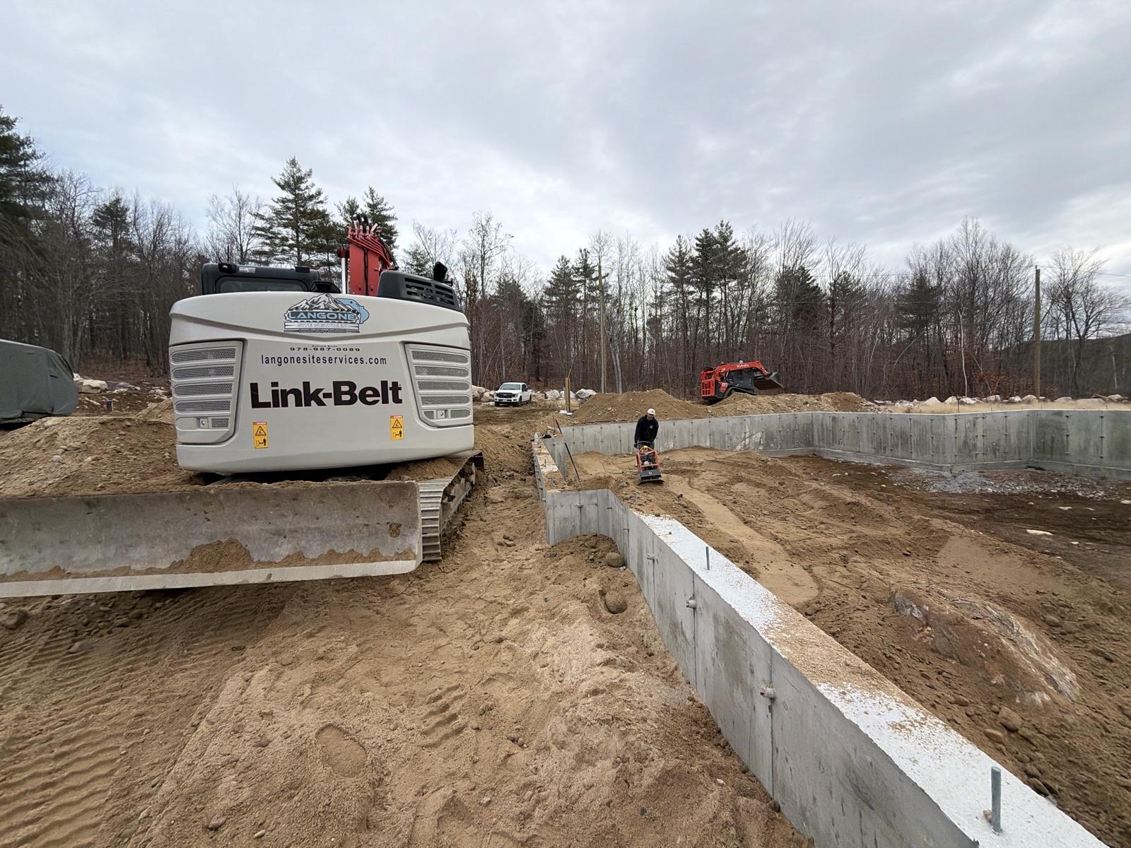 Langone excavator, new foundation, and a crew member compacting fill on a southern Maine lot